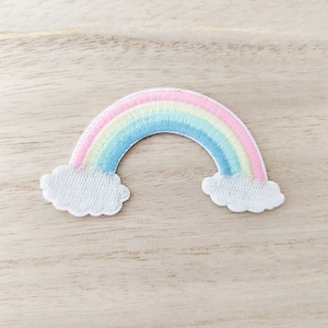May include: Embroidered rainbow patch featuring pastel colors: pink, yellow, and blue. The rainbow arcs over two white cloud shapes. This patch is suitable for clothing, bags, or other fabric items.