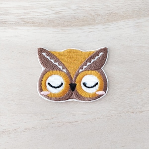 May include: An embroidered owl with closed eyes, featuring brown and yellow stitching. The owl has a white outline and is on a light brown wooden background.