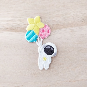 May include: White embroidered patch of an astronaut holding three balloons. The balloons are yellow, blue, and pink with white polka dots.