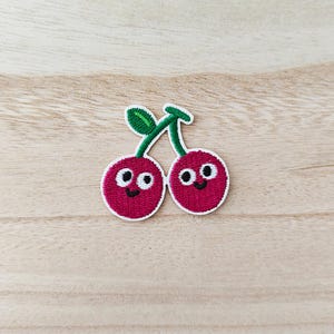 May include: Embroidered patch showing two smiling cherries with white outlines, green stems, and leaves. The cherries are a vibrant red colour with black eyes and smiling mouths. Ideal for adding a touch of fun to clothing or accessories.