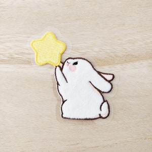 May include: White embroidered bunny holding a yellow star. The bunny is holding the star with its front paws. The bunny is sitting on a clear background.