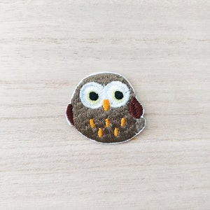 May include: Embroidered owl patch. The patch features a brown body, white and black eyes, and orange accents. It has a white border and is designed for sewing onto fabric. The owl's wings are a darker shade of brown.