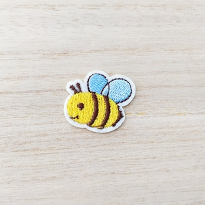 May include: A yellow and brown embroidered bee with blue wings on a white background.
