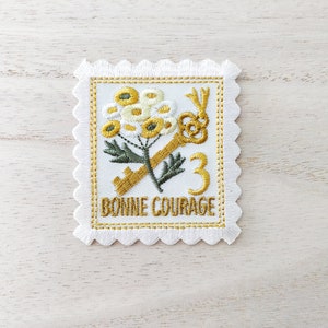 May include: White embroidered patch featuring a yellow flower, a gold key, and the text "Bonne Courage" in gold. The patch has a scalloped edge and a white background.