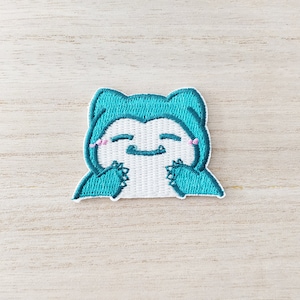 May include: Embroidered patch of a teal cartoon character with a white belly and a smiling face. The character has pink blush marks and is holding its hands up. The patch is designed for application on clothing or accessories.
