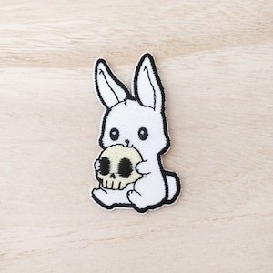 May include: White embroidered patch of a cartoon rabbit clutching a skull. The rabbit has black outlines and details, with black eyes. The skull is beige with black details. The patch is on a light wood grain surface.