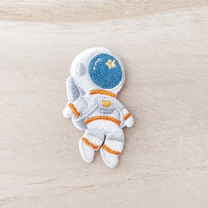 May include: White embroidered astronaut patch with orange details. The astronaut is wearing a helmet with a blue visor and a yellow star.