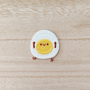 May include: Embroidered patch featuring a smiling fried egg design. The egg has a white outer edge, a yellow yolk with a smiling face, and small red legs and arms. The patch is on a light brown wooden surface.