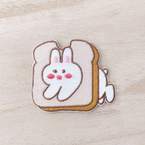May include: An embroidered bunny rabbit peeking out of a brown and beige embroidered slice of bread.