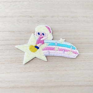 May include: A yellow star with a pink and blue rainbow tail. A girl with blonde hair and a pink dress is sitting on the star.