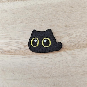 May include: A black embroidered patch shaped like a cat head with large, round eyes. The eyes feature yellow outlines, black pupils, and small white highlights. The patch has a textured surface and a small, rounded tail extension on the right side.