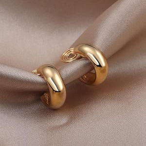 Peut inclure: Une paire de boucles d'oreilles créoles dorées avec une finition lisse et polie. Les boucles d'oreilles présentent un design épais et arrondi et incluent un mécanisme à ressort en spirale à l'arrière, permettant de les porter sans perçage.