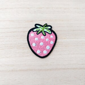 May include: A pink strawberry with white polka dots and a green stem. The strawberry is embroidered on a fabric patch.