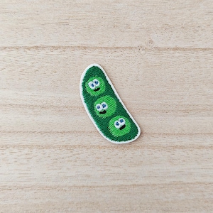 May include: Embroidered patch featuring a green pea pod with three smiling peas. The patch has a white border and is set against a light wood grain background. The peas have blue eyes and are smiling.