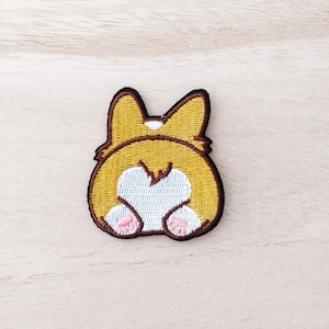 May include: An embroidered patch of a corgi's rear end, featuring brown and white fur. The corgi is sitting with its tail curled up, and the patch has pink paw prints.