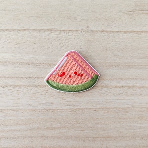 May include: Embroidered patch in the shape of a watermelon slice. The patch features a pink interior, a green rind, and red seed details. The patch is outlined in white and is set against a light wood grain background.