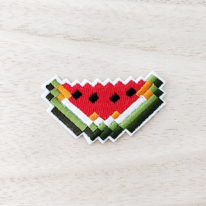 May include: Embroidered watermelon patch with a pixelated design. The patch features a red centre with black seeds, a white rind, and green and orange accents. The patch is on a light wood grain surface.