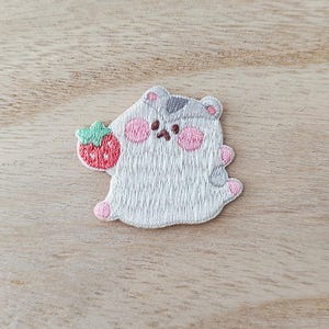 May include: Embroidered patch featuring a cartoon bear holding a strawberry. The bear is white with gray accents, pink cheeks and paws. The strawberry is red with a green stem. The patch is designed for application on clothing or accessories.