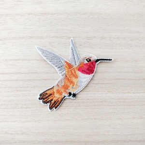May include: Embroidered hummingbird patch with detailed wings and a vibrant colour palette. The bird features a red throat, orange body, and grey wings. The patch is on a light wood grain surface.