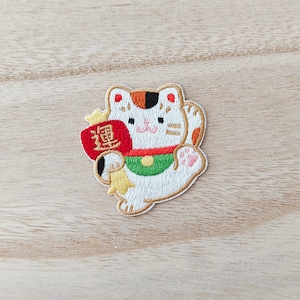 May include: White embroidered patch of a Japanese Maneki-Neko, or lucky cat, with a red and gold coin purse in its left paw and its right paw raised. The cat has orange and black markings on its face and tail. The coin purse has the Chinese character for "good fortune" written on it.
