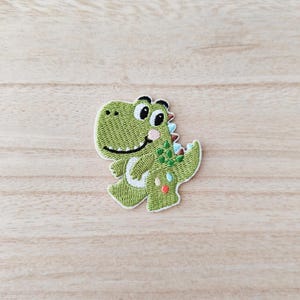 May include: Embroidered patch of a cheerful green dinosaur with a white border. The dinosaur has a smiling face, pink cheeks, and colourful accents on its back and tail. Ideal for adding to clothing or accessories.