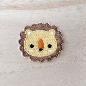 May include: A brown and yellow embroidered lion face patch with a cute expression. The lion has a brown mane and a yellow face with black eyes and a small black nose.