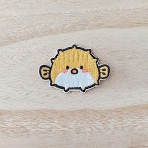 May include: An embroidered patch depicting a cartoon pufferfish. The patch is yellow with a spiky outline, small black eyes, and rosy cheeks. The pufferfish has small fins and a white belly. The patch is designed for clothing.