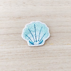 May include: A light blue embroidered seashell patch with a white border. The seashell design features detailed stitching in varying shades of blue, creating a textured appearance. The patch is likely for decorative purposes, such as on clothing or accessories.
