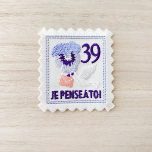 Peut inclure: Un patch brodé blanc en forme de timbre-poste avec une pensée violette, une colombe blanche portant une enveloppe rose et le texte "JE PENSE A TOI" et "39".