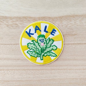 May include: A round, white embroidered patch with a yellow sunburst design and the word "KALE" in blue letters. A green leafy vegetable with a pink heart and a smiling face is in the center of the patch.