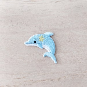 May include: A light blue embroidered dolphin with a yellow star on its back. The dolphin is leaping out of the water.