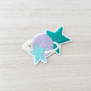 May include: Purple and teal embroidered patch featuring a planet with a ring and a star inside, along with two stars. The patch is ideal for adding a touch of whimsy to your clothing or accessories.