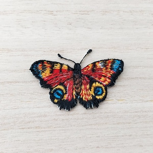 May include: Embroidered butterfly patch with red, yellow, black and blue details. The butterfly has a brown body and black antennae.