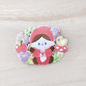 May include: Embroidered iron-on patch featuring a cartoon girl in a red hooded dress, surrounded by flowers, an apple, and a mushroom with a ladybug on top.