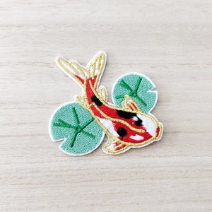 May include: Embroidered patch of a koi carp in red, black, and white, with gold detailing. The fish is above two green lily pads. The patch has a white border and is suitable for clothing or accessories.