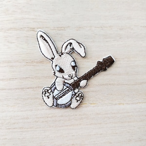 Peut inclure: Écusson brodé représentant un lapin de dessin animé jouant de la guitare. Le lapin est gris clair avec des yeux bleus et tient une guitare marron. L'écusson a une bordure blanche et est sur un fond de grain de bois clair.