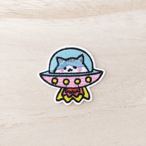 May include: Embroidered iron-on patch featuring a cartoon cat in a pink and blue spaceship with yellow accents. The cat is looking out of the spaceship window.