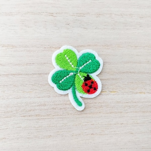 May include: Embroidered four-leaf clover patch with a white border. The clover is green with white stitching and a red and black ladybug on one leaf. The patch is on a light wood grain surface.