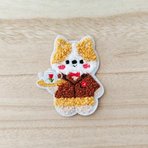 May include: Embroidered patch of a cartoon corgi in a brown suit and red bow tie, holding a plate with a rose. The patch has a white background and a light brown border. The corgi's fur is a mix of white, brown, and yellow.