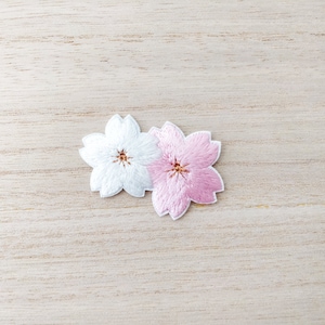 May include: Two embroidered cherry blossom flowers, one white and one pink, on a light wood background.