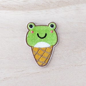 May include: Embroidered patch of a green frog ice cream cone with a smiling face and pink cheeks.