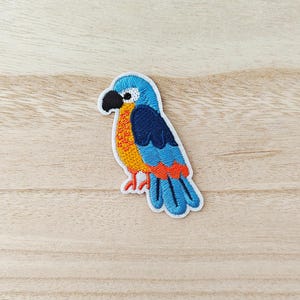 May include: Embroidered patch of a colorful parrot. The bird features a blue head and wings, orange chest, and red accents. It has a black beak and is outlined in white. The patch is designed to be ironed on or sewn onto fabric.