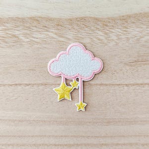 May include: Embroidered patch featuring a white cloud with a pink outline. Two yellow stars hang below the cloud, connected by pink lines. The patch is on a light brown wooden surface.