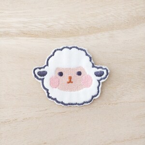 May include: A white embroidered sheep face patch with pink cheeks and a black outline.