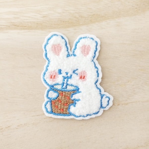 Peut inclure: Un lapin blanc brodé tenant une tasse brune avec une paille rose. Le lapin a des oreilles et des joues roses. Le patch brodé est sur un fond de bois brun clair.