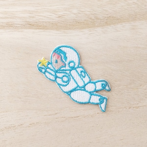 Peut inclure: Patch brodé blanc représentant un astronaute avec des contours bleus, tenant une étoile jaune. L'astronaute porte un casque et a un visage rose.