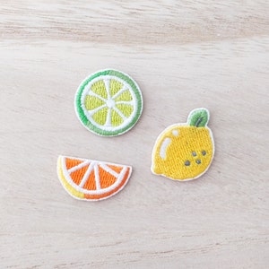 May include: Three embroidered fruit patches: a lime slice, an orange slice, and a lemon. The lime is green and white, the orange is orange and white, and the lemon is yellow with a green leaf.