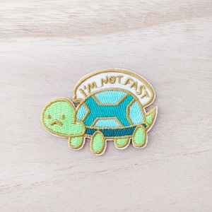 May include: A green and blue embroidered patch of a turtle with the text "I'm not fast" on a white background.