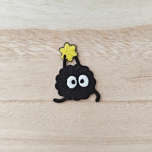 May include: A black embroidered patch of a cartoon character with large white eyes and a small yellow star held above its head. The character has a fluffy, round body and small, curved legs. The patch is on a light brown wooden surface.