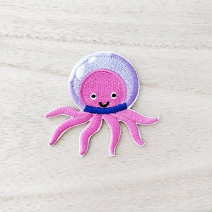 May include: Pink embroidered octopus with a purple and white bubble helmet and a blue ribbon around its head.